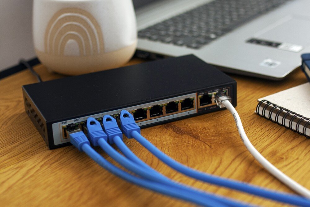 Switch EXTRALINK Perses EX-SG1008PE Czarny przełącznik sieciowy na drewnianym biurku z podłączonymi czterema niebieskimi kablami i jednym białym, w tle widać laptopa i kubek, porty RJ-45, przepustowość, algorytm przełączania