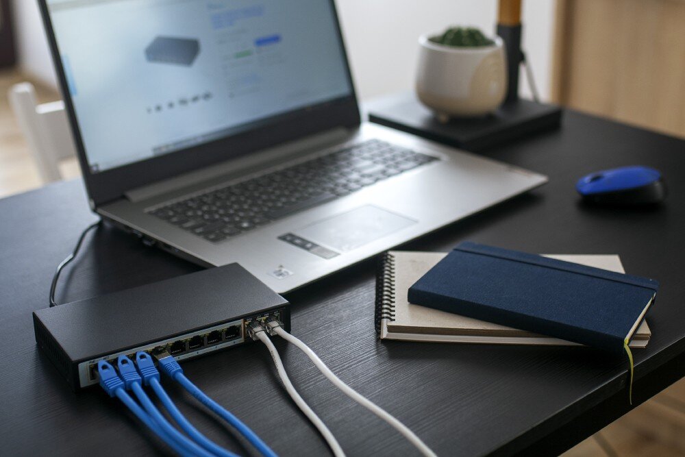 Switch EXTRALINK Perses EX-SG1008PE Czarny przełącznik sieciowy na czarnym biurku obok otwartego laptopa, myszki i notatników, do przełącznika podłączone są cztery niebieskie i dwa białe kable, standardy, zabezpieczenia