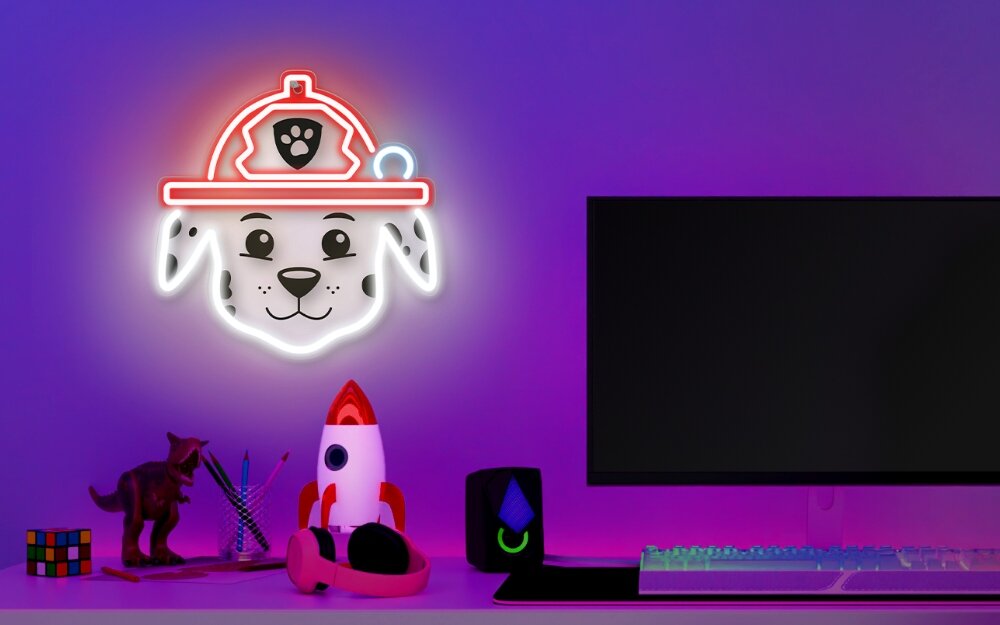 Na ścianie wisi neon LED przedstawiający twarz psa z czarnymi uszami i nosem, w czerwonym hełmie z symbolem łapy. Obok znajduje się biurko z monitorem, klawiaturą, figurką rakiety i innymi drobnymi przedmiotami.