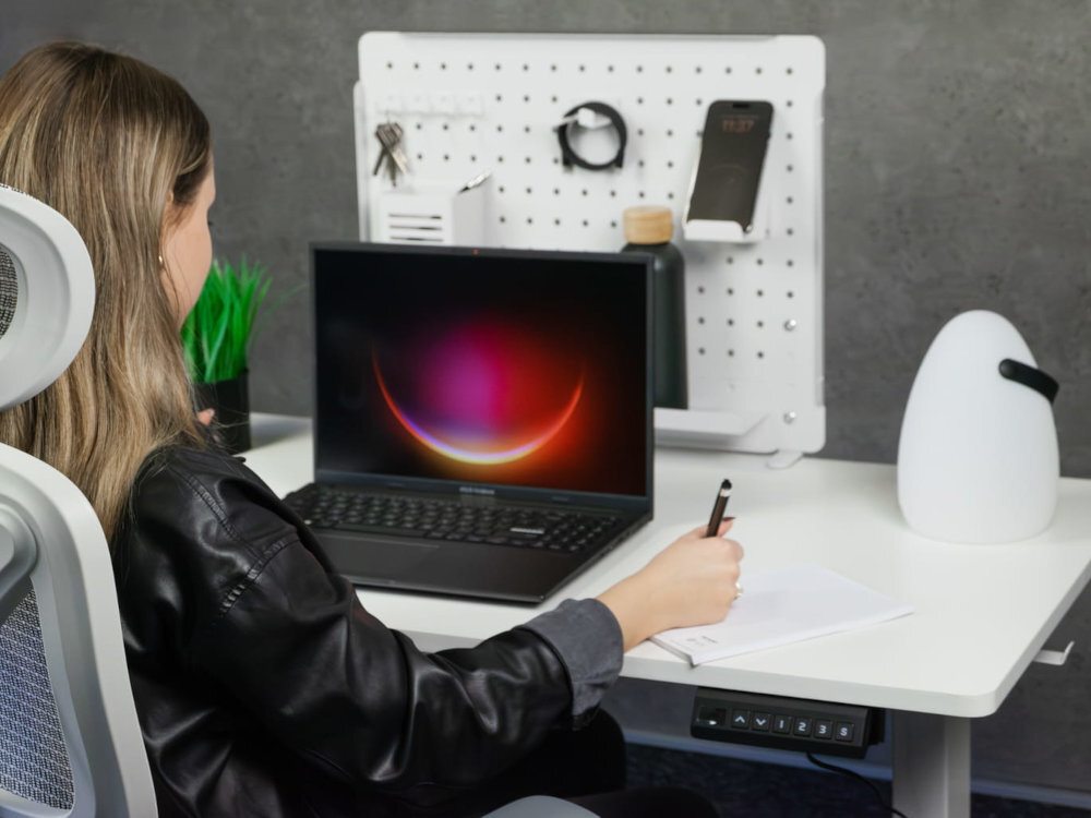 Osoba siedzi przy białym biurku, na którym znajduje się laptop z kolorowym ekranem, notatnik oraz lampka. Na ścianie widać panel z uchwytami na akcesoria i telefon.
