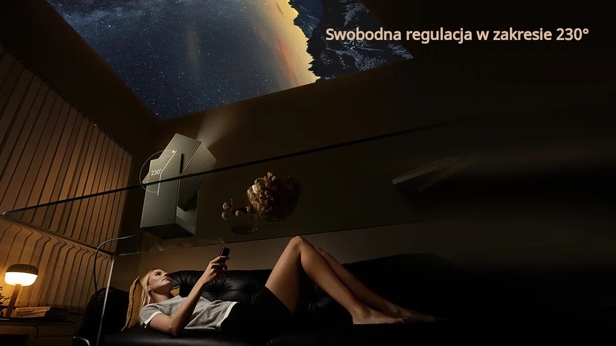 Osoba leży na kanapie, trzymając pilot, a na suficie wyświetlany jest obraz z projektora. Widoczny jest tekst: \'Swobodna regulacja w zakresie 230°\'.