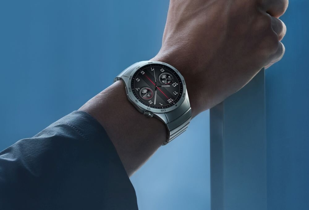 Osoba nosi smartwatch z metalową bransoletą i okrągłą tarczą z trzema małymi cyferblatami. Tło jest jednolite, w odcieniach niebieskiego.