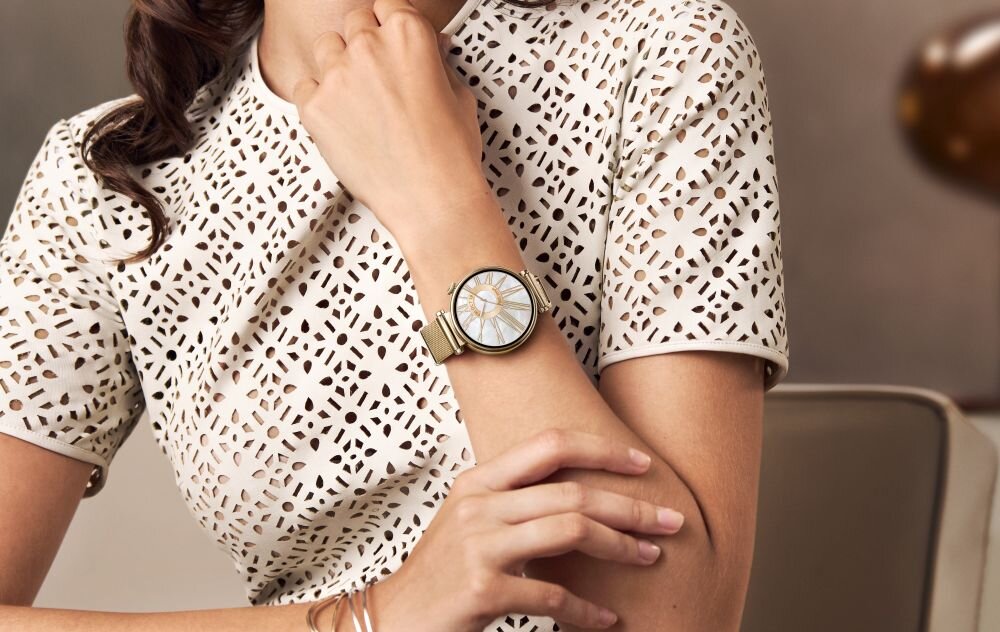 Osoba w białej, ażurowej bluzce prezentuje smartwatch na nadgarstku. Zegarek ma okrągłą tarczę z jasnym wyświetlaczem i metalową bransoletą.