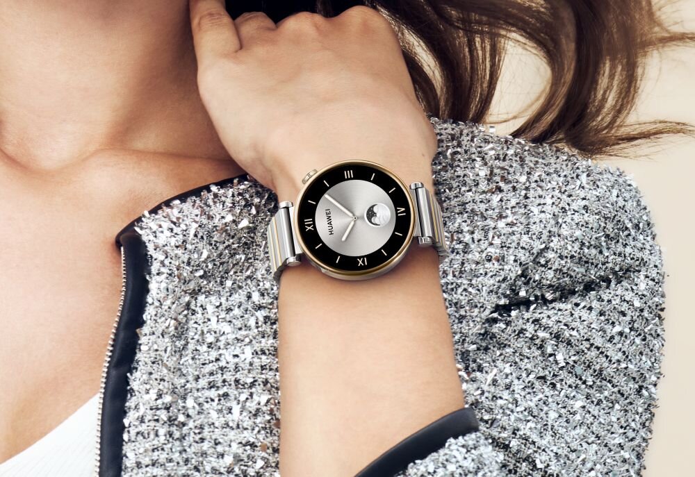 Osoba nosi smartwatch z okrągłą tarczą, na której widnieje napis \'HUAWEI\'. Zegarek ma srebrną bransoletę i czarną tarczę z rzymskimi cyframi.