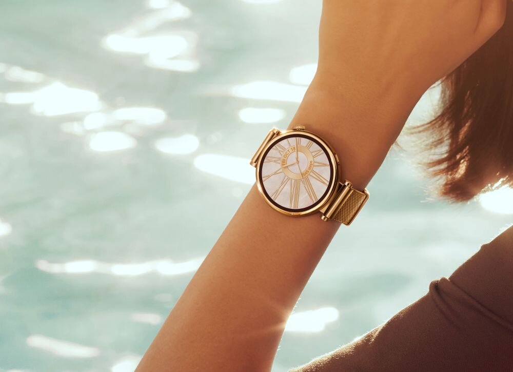 Na zdjęciu widoczny jest smartwatch z okrągłą tarczą i złotą bransoletą, noszony na nadgarstku. W tle znajduje się rozmyty obraz wody.
