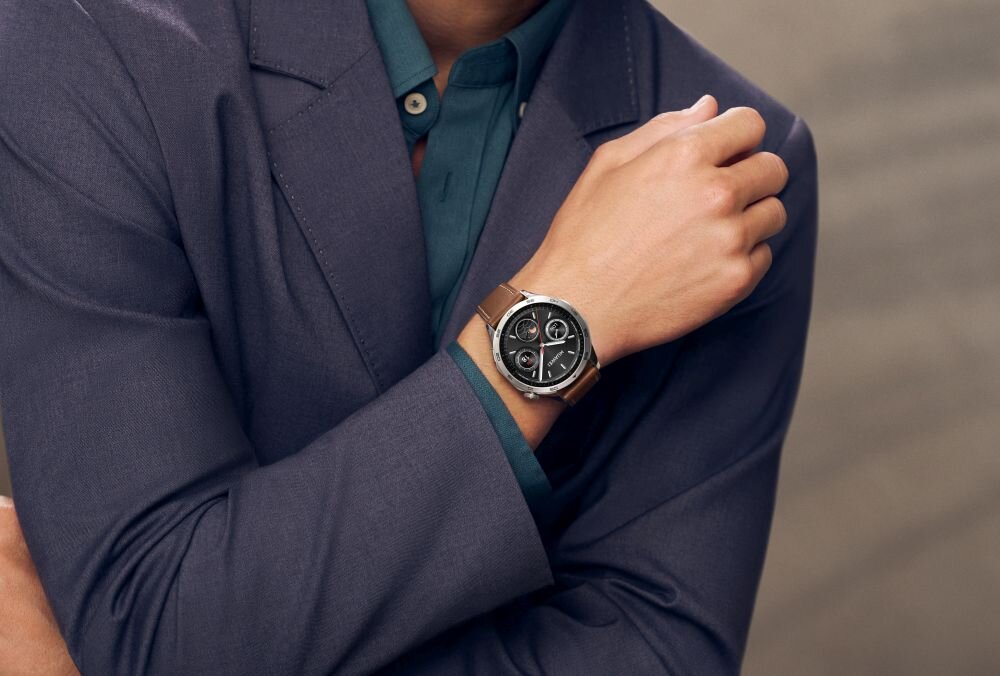 Osoba w eleganckiej marynarce prezentuje smartwatch na nadgarstku. Zegarek ma okrągłą tarczę i brązowy pasek.