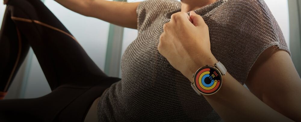 Osoba siedzi, nosząc smartwatch z okrągłym, kolorowym wyświetlaczem. Na ekranie zegarka widoczne są koncentryczne okręgi w różnych kolorach.