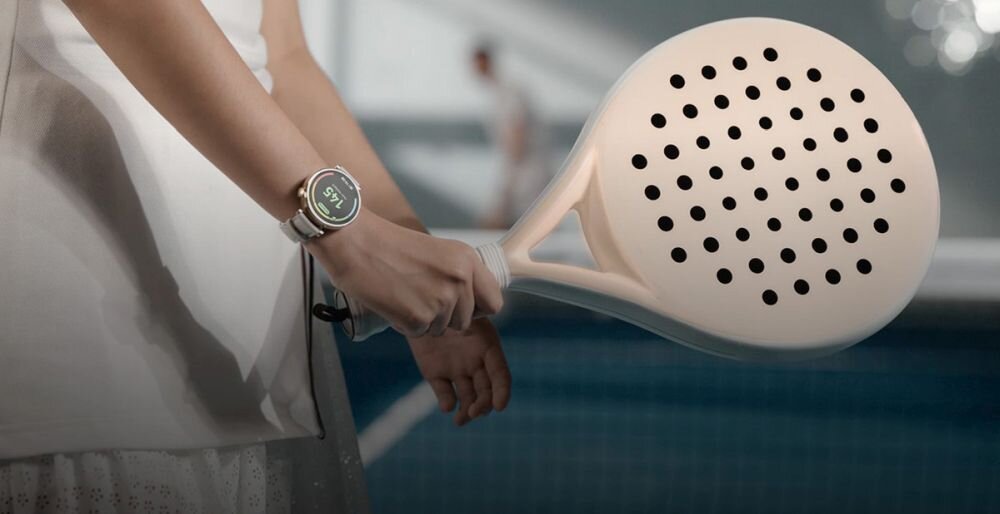 Osoba trzyma rakietę do padla, na nadgarstku widoczny jest smartwatch z okrągłym wyświetlaczem. W tle rozmyta postać na korcie tenisowym.