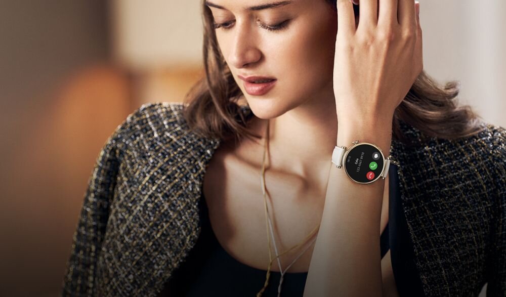 Osoba nosi smartwatch z okrągłym wyświetlaczem, na którym widoczne są ikony połączenia i tekst. Smartwatch ma biały pasek i metalową kopertę.