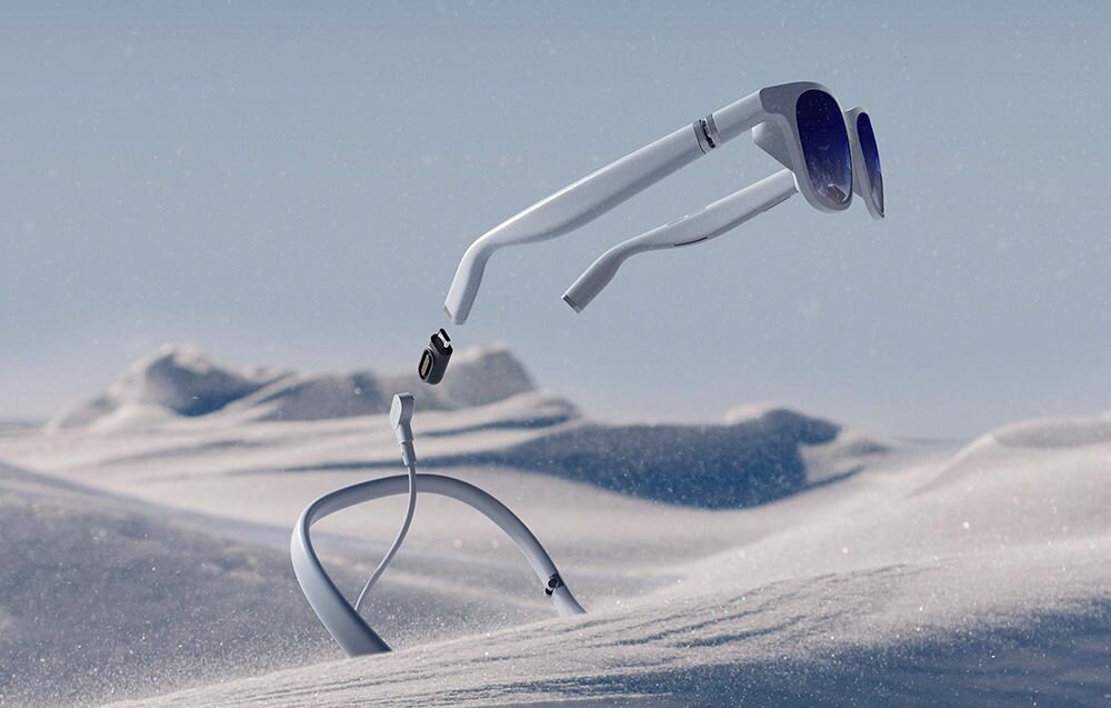 Na obrazie widać białe okulary przeciwsłoneczne unoszące się nad zaśnieżonym krajobrazem. Obok okularów znajduje się złącze magnetyczne do adaptera USB-C.