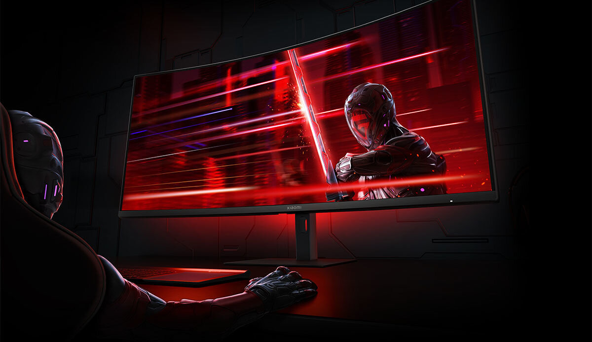 Na obrazie widoczny jest zakrzywiony monitor z dynamiczną grafiką przedstawiającą postać w futurystycznym stroju. Na dolnej ramce monitora znajduje się logo \'XIAOMI\'.