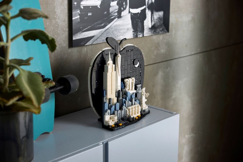Na szafce stoi model LEGO przedstawiający panoramę miasta w kształcie jabłka. W tle widać czarno-białe zdjęcie ludzi na ulicy.