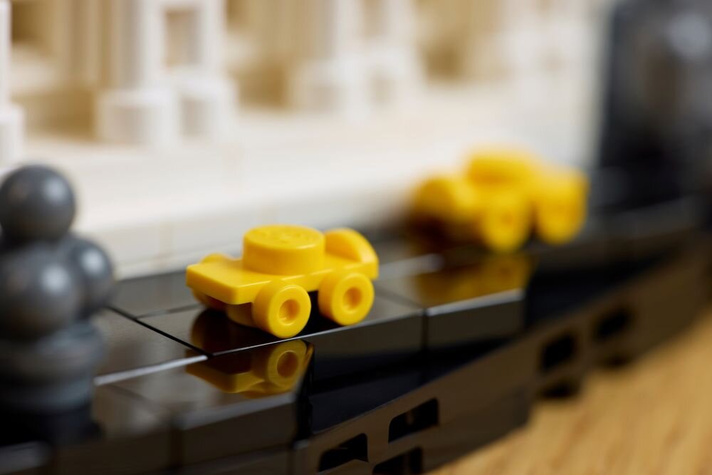 Na obrazie widoczne są małe, żółte modele samochodów z klocków LEGO ustawione na czarnej podstawie. W tle znajduje się fragment białej konstrukcji z klocków.