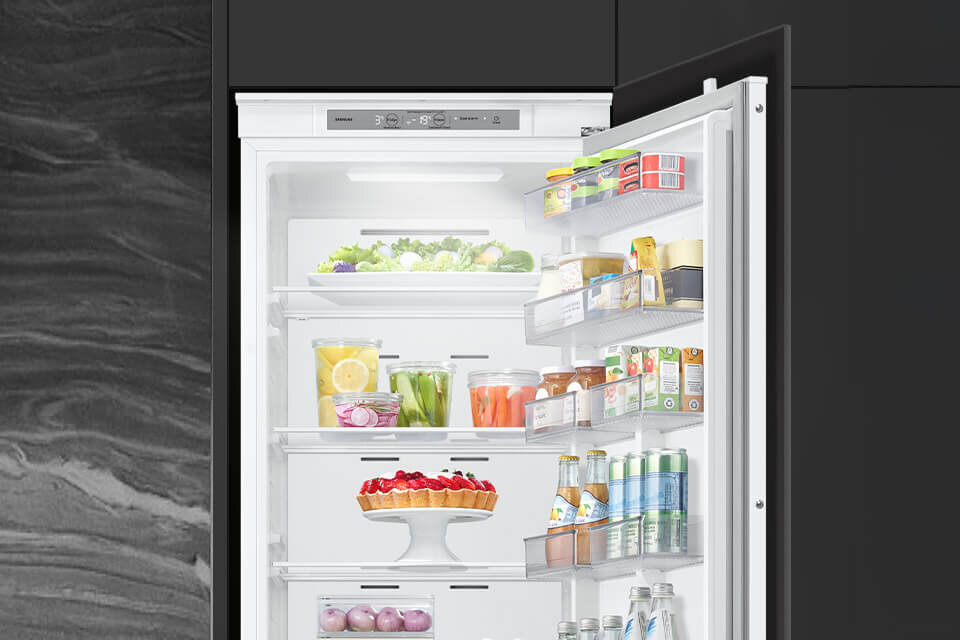 Otwarta lodówka z widocznymi półkami wypełnionymi różnorodnymi produktami spożywczymi, takimi jak warzywa, napoje i ciasta. Na drzwiach znajdują się dodatkowe półki z produktami w słoikach i kartonach.