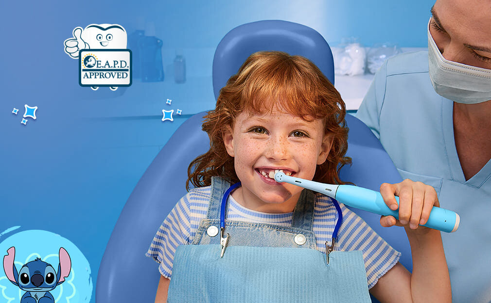 Dziecko siedzi na fotelu dentystycznym, trzymając szczoteczkę magnetyczną ORAL-B IO w ustach. Obok widoczna jest postać z kreskówki oraz logo z napisem \'E.A.P.D. APPROVED\'.