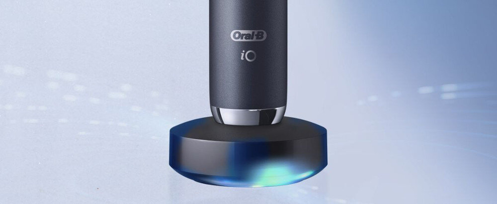 Czarna szczoteczka magnetyczna Oral-B iO stoi na okrągłej podstawce z niebieskim podświetleniem. Na szczoteczce widoczne jest logo \'Oral-B iO\'.