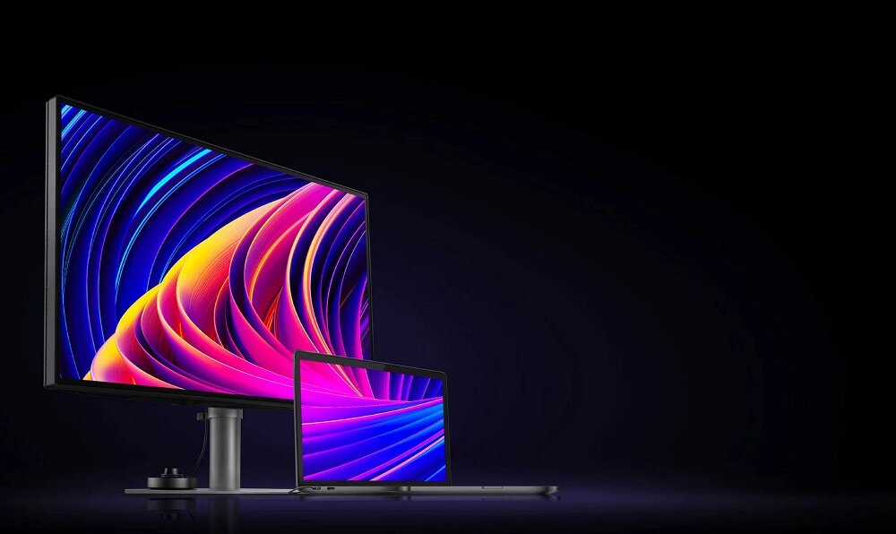 Na obrazie widoczny jest duży monitor oraz laptop, oba wyświetlają kolorową, abstrakcyjną grafikę z falistymi wzorami. Tło jest ciemne, co podkreśla jasność i intensywność kolorów na ekranach.