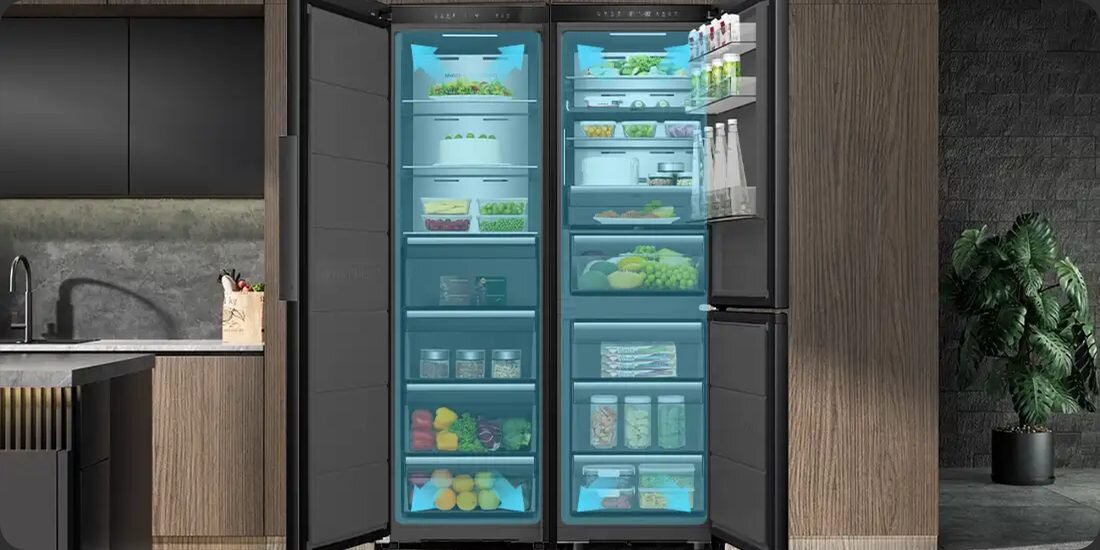 Otwarta lodówka z widocznymi półkami wypełnionymi różnorodnymi produktami spożywczymi, takimi jak warzywa, owoce i napoje. Wnętrze lodówki jest oświetlone na niebiesko, a obok znajduje się nowoczesna kuchnia z drewnianymi elementami.