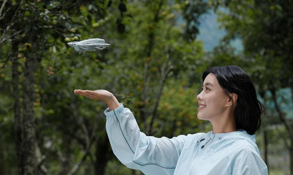 Osoba w jasnej kurtce stoi na tle drzew, trzymając rękę w górze, a nad nią unosi się mały dron.