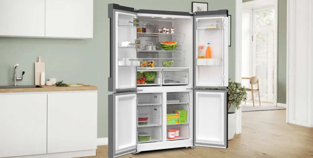 Otwarta lodówka z czterema drzwiami, wypełniona różnymi produktami spożywczymi, takimi jak warzywa, napoje i pojemniki z jedzeniem. W tle widoczna jest kuchnia z jasnymi szafkami i drewnianym blatem.