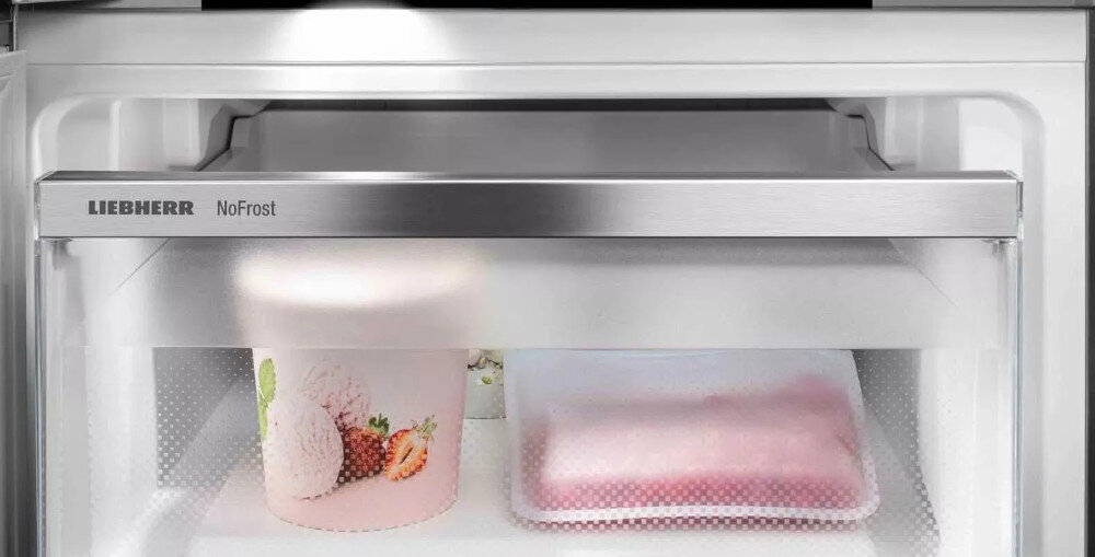 Wnętrze zamrażarki z widocznym pojemnikiem na lody z owocami na etykiecie i zapakowanym produktem spożywczym. Na górnej krawędzi widnieje napis \'LIEBHERR NoFrost\'.