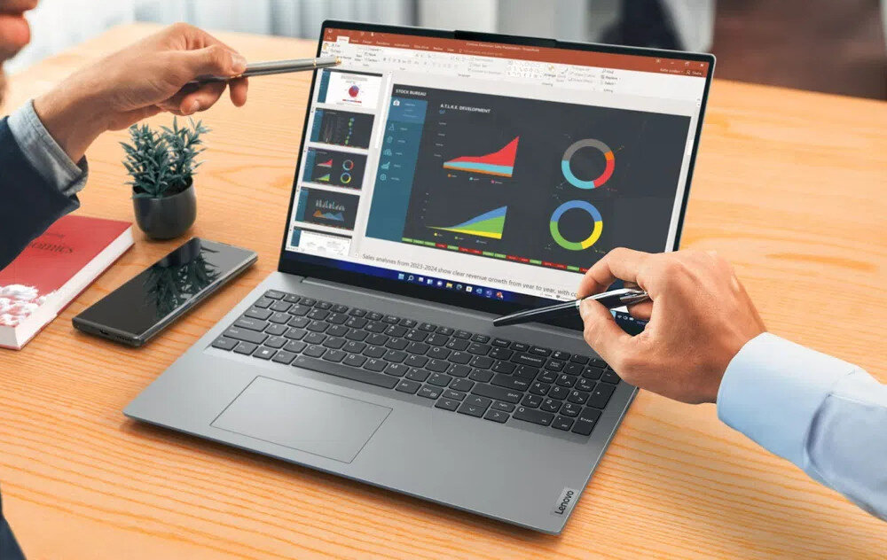 Na drewnianym stole znajduje się otwarty laptop z widocznym ekranem, na którym wyświetlane są wykresy i prezentacja. Obok laptopa leży smartfon, a dwie osoby wskazują na ekran długopisami.