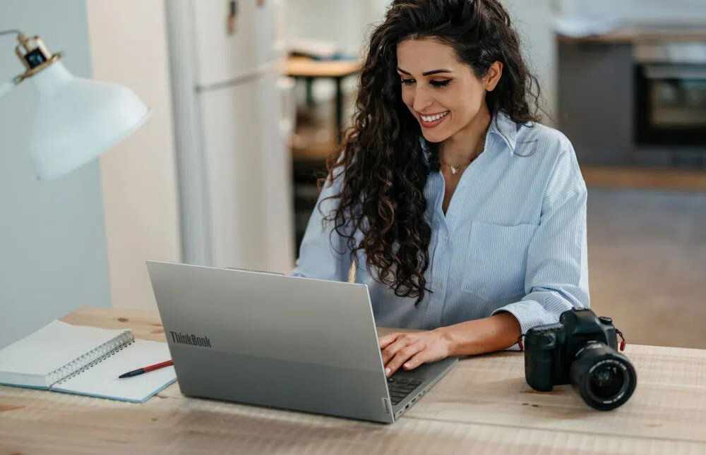 Osoba siedzi przy stole i korzysta z laptopa marki ThinkBook. Obok znajduje się aparat fotograficzny i notatnik.