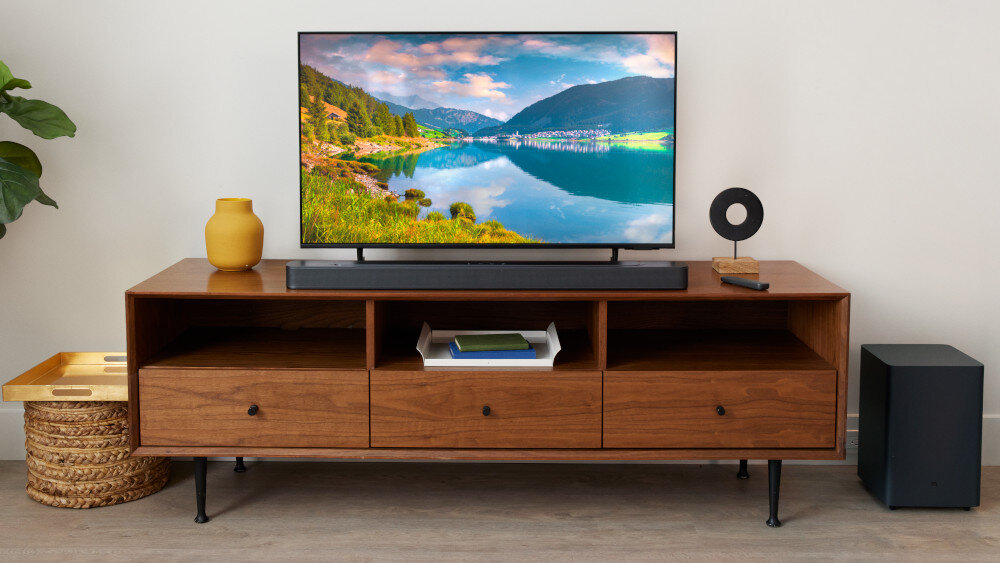 Na drewnianej szafce stoi telewizor z krajobrazem jeziora i gór na ekranie. Przed telewizorem znajduje się soundbar, a obok szafki stoi subwoofer.