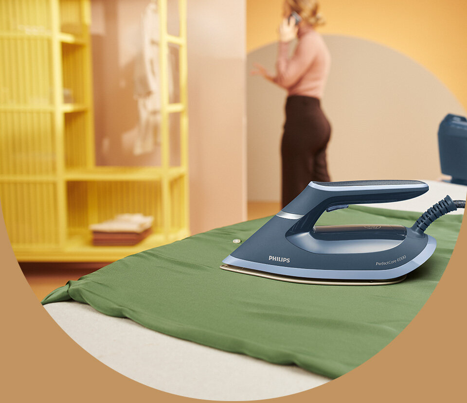 Na zielonym materiale leży niebieskie żelazko z napisem \'PHILIPS PerfectCare 6000\'. W tle widoczna jest osoba rozmawiająca przez telefon oraz żółta półka.