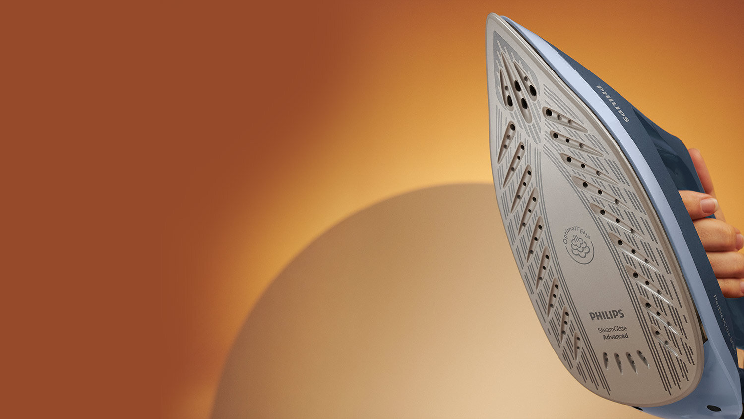 Zdjęcie przedstawia stopę żelazka z widocznymi otworami parowymi i napisem \'PHILIPS SteamGlide Advanced\'. W tle znajduje się rozmyte, pomarańczowe tło.
