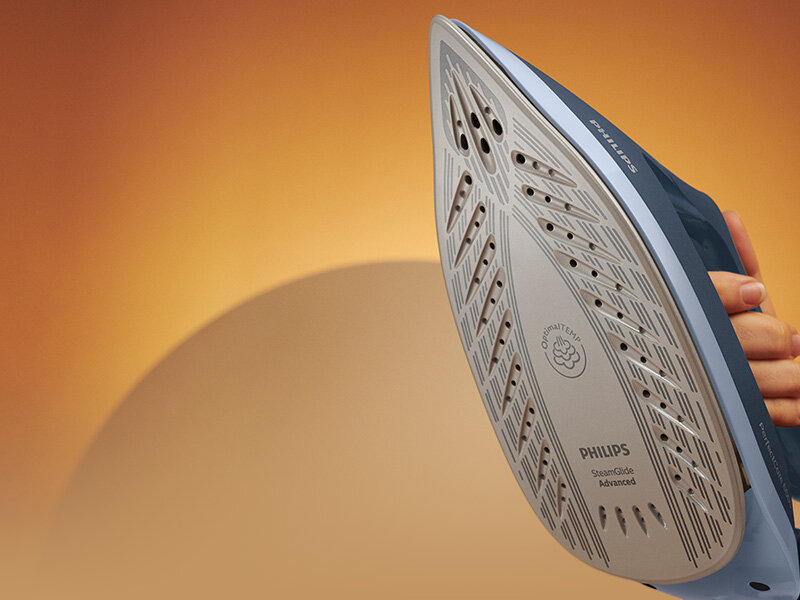 Zdjęcie przedstawia stopę żelazka z widocznymi otworami parowymi i napisem \'PHILIPS SteamGlide Advanced\'. W tle znajduje się gradientowe tło w odcieniach pomarańczowych.