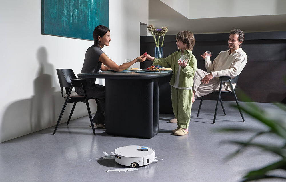 Na podłodze w jasnym pomieszczeniu znajduje się biały robot sprzątający. W tle przy stole siedzą trzy osoby, które jedzą posiłek.