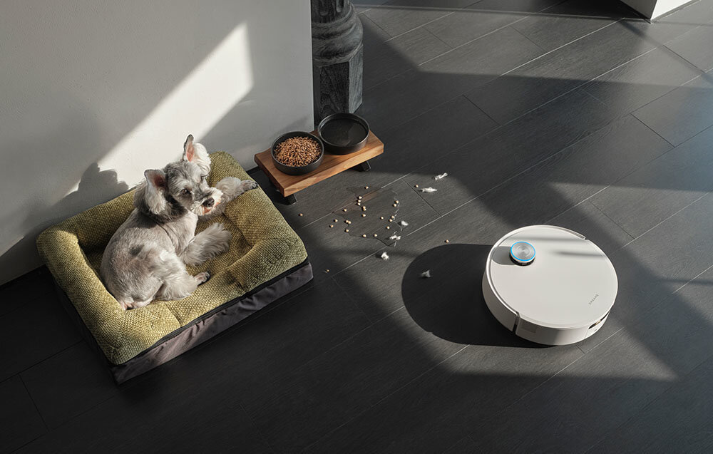 Na podłodze znajduje się biały robot sprzątający obok miski z jedzeniem i legowiska z psem. Wokół robota widoczne są rozsypane drobinki jedzenia.