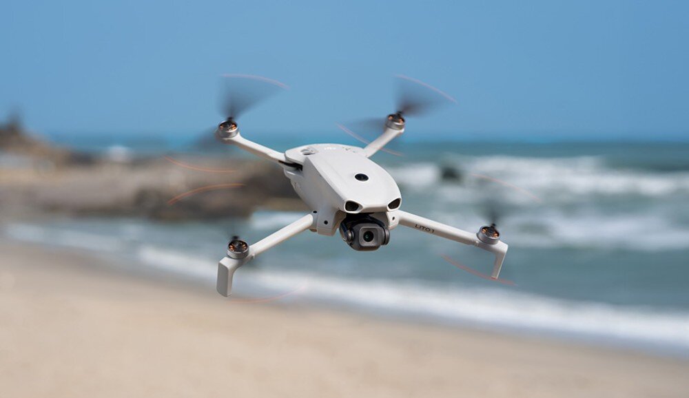 Na obrazie widoczny jest biały dron w locie nad plażą z falującym morzem w tle. Dron ma cztery wirniki i kamerę z przodu.
