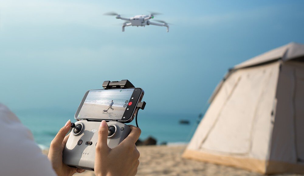 Osoba trzyma kontroler z zamocowanym smartfonem, na ekranie którego widać obraz z kamery drona. W tle znajduje się plaża, namiot i latający dron.
