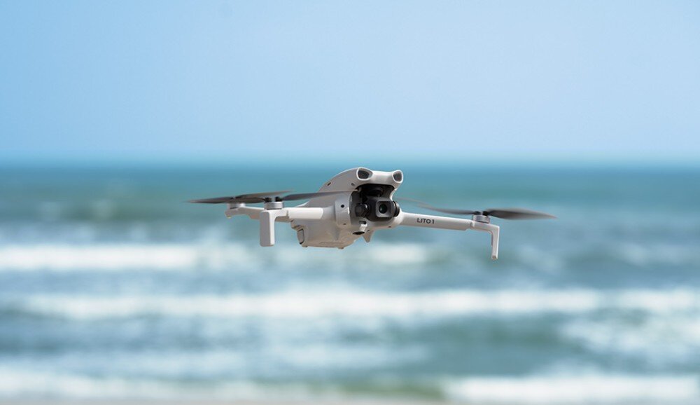 Na obrazie widoczny jest biały dron w locie nad morzem. W tle widać rozmyte fale i niebo.