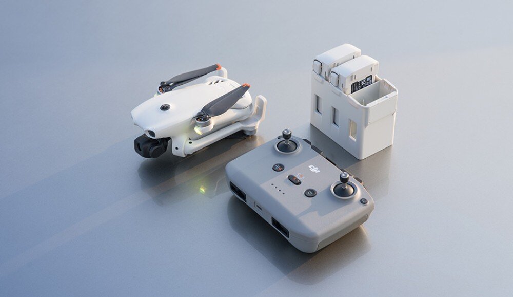 Na obrazie znajduje się biały dron z złożonymi śmigłami, szary kontroler z logo DJI oraz stacja dokująca z dwoma bateriami. Wszystkie elementy są umieszczone na gładkiej, jasnej powierzchni.