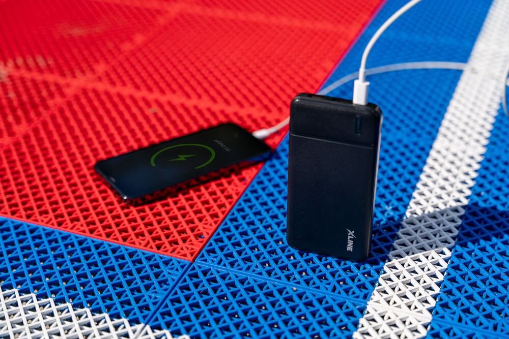 Czarny powerbank z logo \'XLINE\' ładuje smartfon, który pokazuje ikonę ładowania. Oba urządzenia znajdują się na kolorowej, perforowanej powierzchni w kolorach czerwonym, niebieskim i białym.