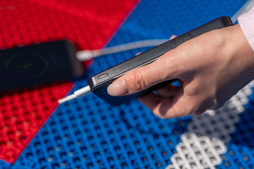 Osoba trzyma czarny powerbank z podłączonym kablem na tle kolorowej powierzchni z czerwonymi, niebieskimi i białymi elementami. W tle widać inny czarny przedmiot z zielonym symbolem błyskawicy.