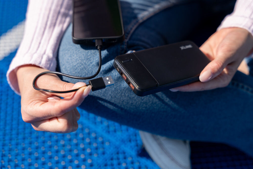 Osoba trzyma czarny powerbank z logo XLINE, podłączony do smartfona za pomocą kabla USB. Powerbank ma dwa widoczne porty USB i jest używany na tle niebieskiej powierzchni.