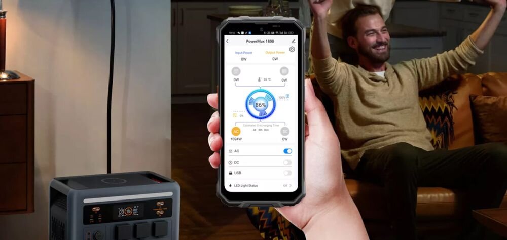 Osoba trzyma smartfon z aplikacją pokazującą stan naładowania stacji zasilania PowerMax 1800 na poziomie 86%. W tle widać stację zasilania oraz osobę siedzącą na kanapie z uniesionymi rękami.