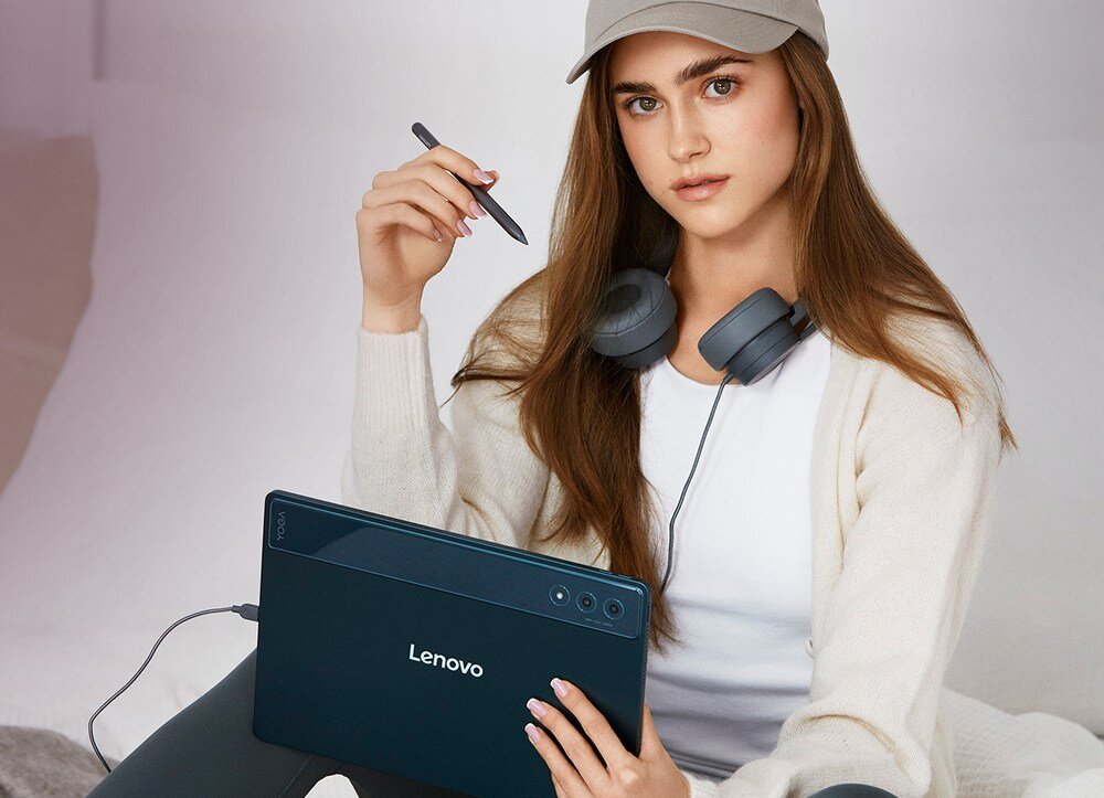 Osoba trzyma tablet marki Lenovo w kolorze niebieskim, z widocznym logo \'Lenovo\' na tylnej obudowie. W ręku trzyma rysik, a na szyi ma zawieszone słuchawki.