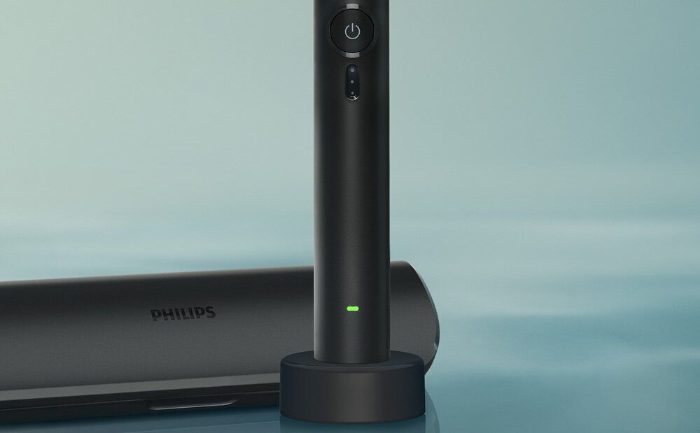 Czarna szczoteczka soniczna na stojaku z widocznym przyciskiem zasilania i zielonym wskaźnikiem. W tle znajduje się etui z napisem \'PHILIPS\'.