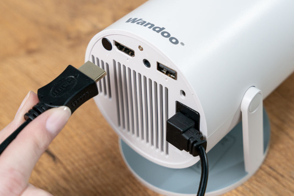 Biały projektor z logo \'Wandoo\' na górze, z widocznymi portami HDMI i USB. Osoba podłącza kabel HDMI do jednego z portów.
