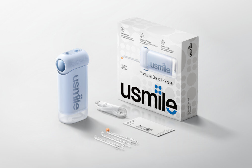 Na obrazie znajduje się niebieski irygator z napisem \'usmile\' oraz pudełko z napisem \'Portable Dental Flosser\'. Obok leżą akcesoria, w tym końcówki i kabel, a także instrukcja obsługi.
