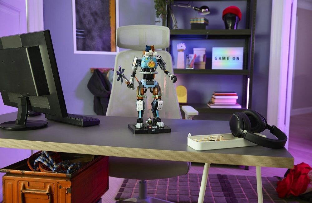 Na biurku znajduje się figurka robota z klocków LEGO oraz monitor, słuchawki i pudełko. W tle na półce widoczny jest napis \'GAME ON\'.