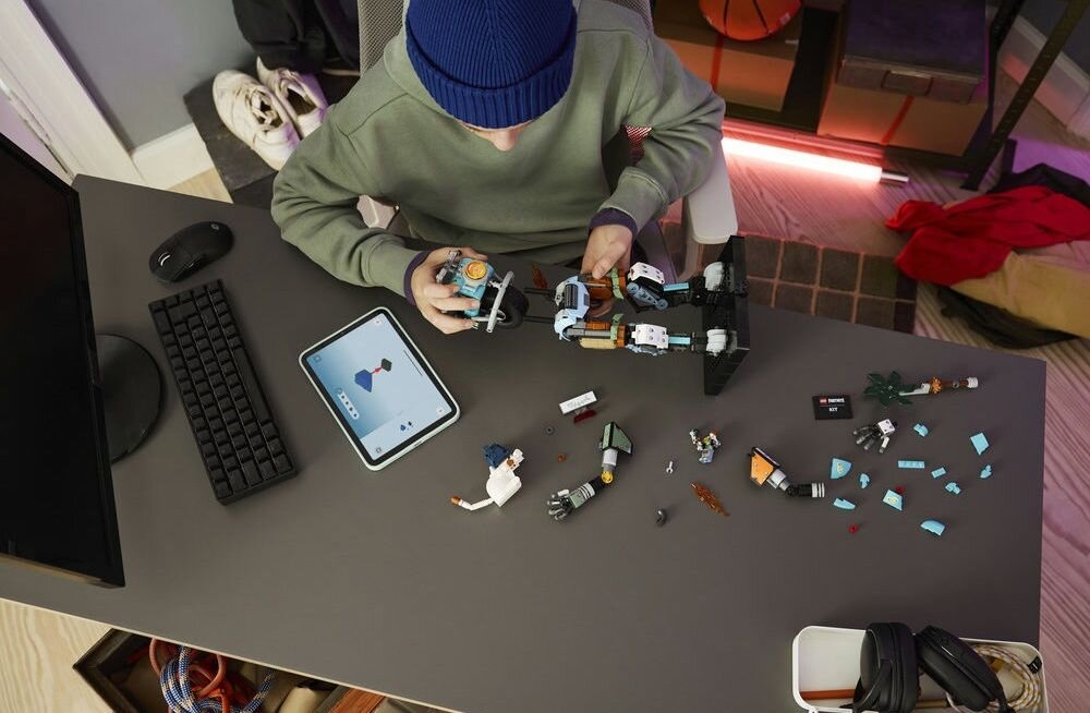 Osoba siedzi przy biurku, składając zestaw klocków LEGO. Na biurku znajduje się tablet z wyświetlonym obrazem oraz rozłożone elementy klocków.