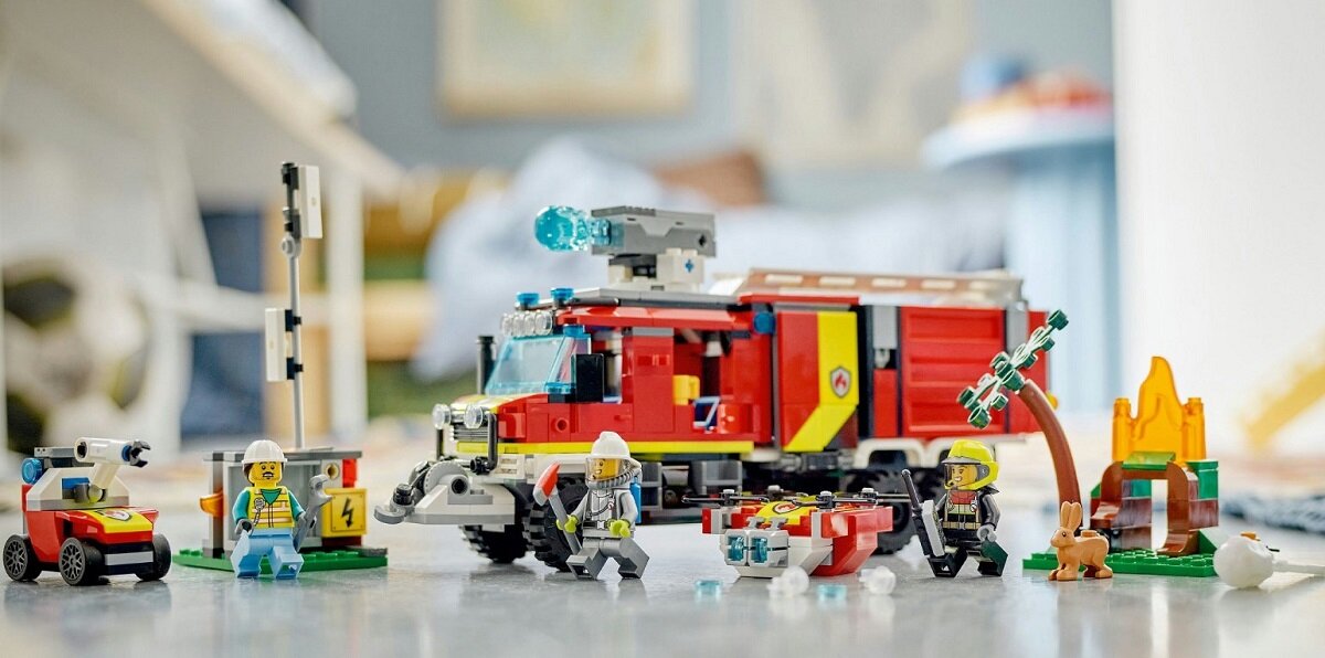 Zestaw klocków LEGO przedstawiający czerwony terenowy pojazd straży pożarnej z wysięgnikiem, otoczony figurkami strażaków i akcesoriami, takimi jak drzewo i płomienie. W tle widoczne są rozmyte elementy wnętrza pokoju.