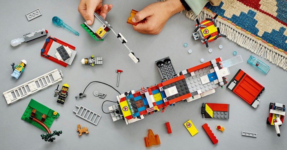 Na stole rozłożone są elementy zestawu LEGO, w tym części pojazdu straży pożarnej, figurki strażaków i akcesoria. Dwie osoby składają model z klocków LEGO.