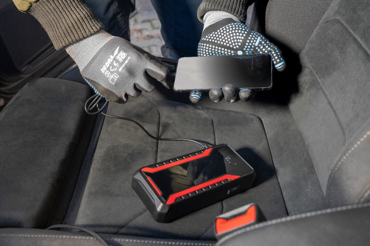 Na siedzeniu samochodowym leży czarny powerbank z czerwonymi akcentami, podłączony do smartfona trzymanego przez osobę w rękawiczkach. Na rękawiczkach widoczny jest napis \'DEDRA\'.